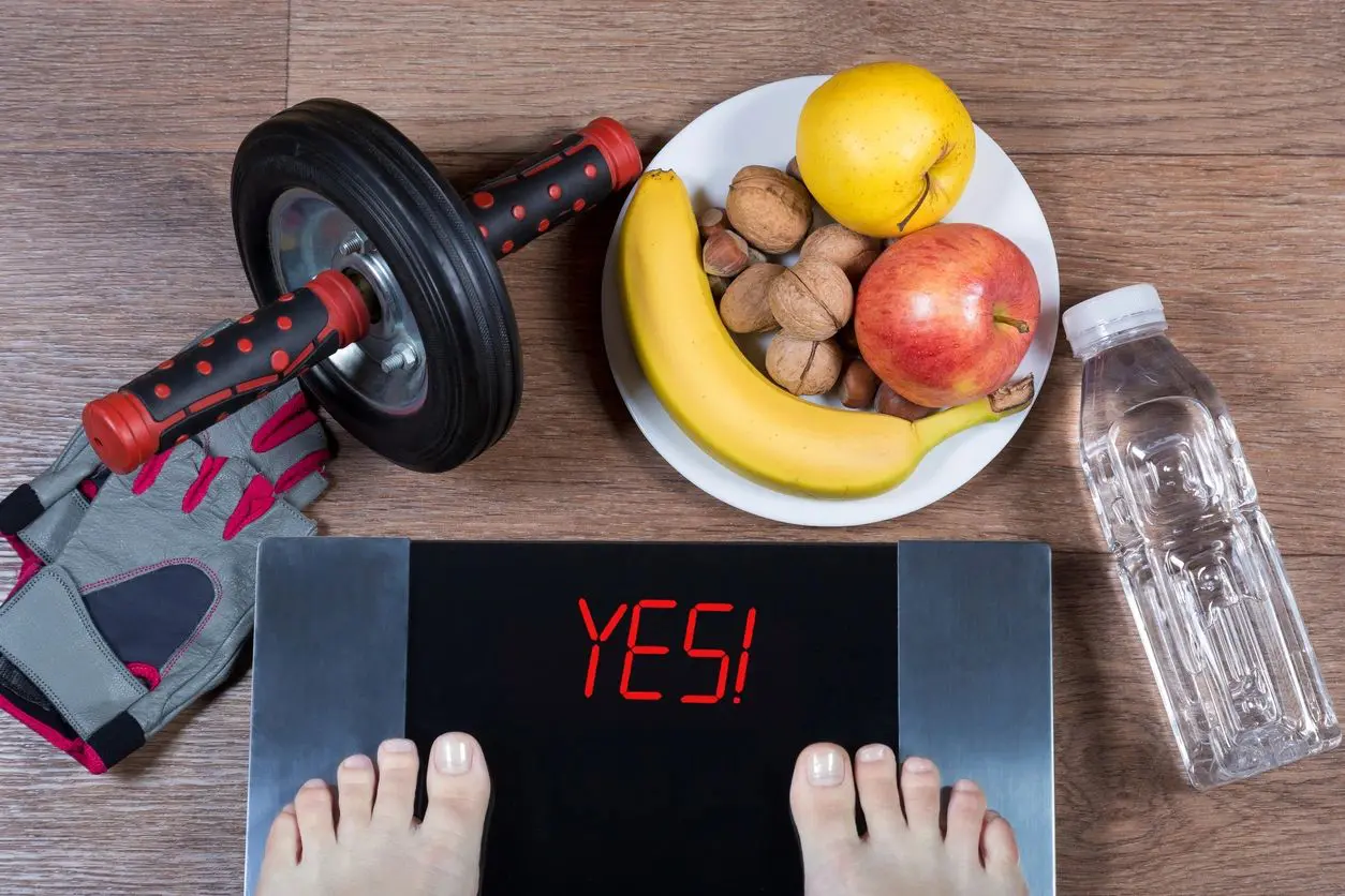 A person standing on their scale with an apple, banana and other fruits in front of them.